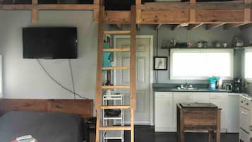 Habitación estilo loft con cama, una escalera que lleva a una zona elevada, cocina y televisor montado en la pared.