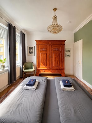 Une chambre d’hôtel avec deux lits, une armoire en bois, un fauteuil vert, un lustre et une fenêtre avec des rideaux.