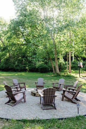 A circular seating area with Adirondack chairs around a central table, set on a gravel surface with trees in the background.