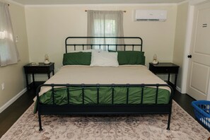 Ein Bett mit Metallgestell, Bettwäsche in Grün und Weiß, zwei Nachttische mit Lampen, eine Wand-Klimaanlage und ein gemustertes Teppich.