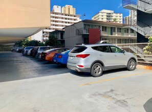 Un estacionamiento con varios coches, incluyendo un SUV blanco, y un edificio de varios pisos con balcones al fondo.