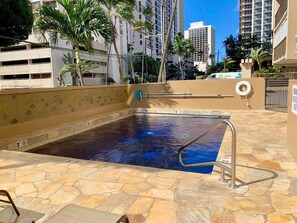 A swimming pool with a stone deck, surrounded by palm trees and high-rise buildings. - Waikiki Getaway | 2 King Beds, Pool & Full Kitchen (Honolulu)