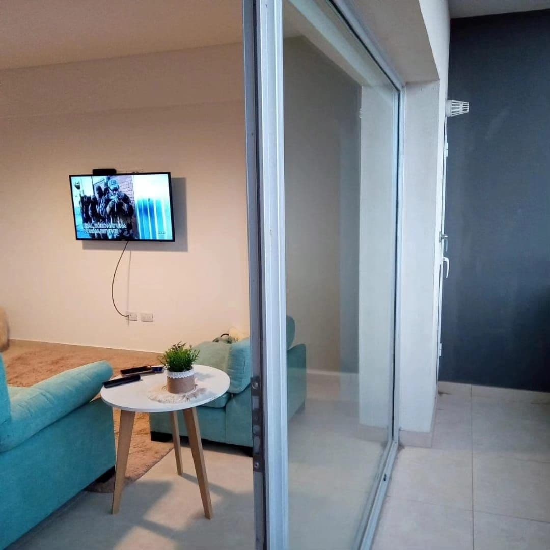 Una sala de estar moderna con un televisor de pantalla plana montado en la pared, un sofá color turquesa, una pequeña mesa redonda con una planta y un ventanal corredizo que da a un balcón.