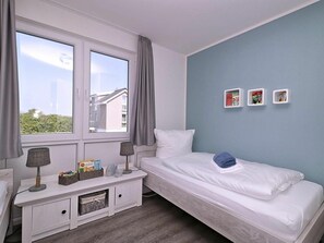 A bedroom with a single bed, a bedside table with lamps, a window with curtains, and a wall with picture frames. - House No. 34, Holiday Home Fischerhus (Wangerland)