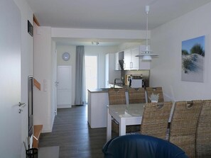 Una cocina moderna con armarios blancos, un comedor con sillas de mimbre y vistas al exterior a través de una ventana.