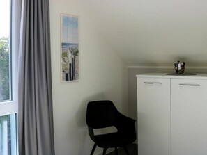 Una habitación con ventana, una silla, un armario blanco y un cuadro en la pared.