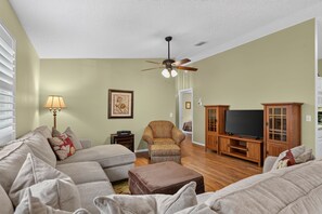 A living room with a sofa, ottoman, armchair, ceiling fan, television, and wooden entertainment center. - 2472 Z3A Village of Lynnhaven (The Villages)