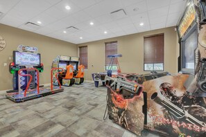 A gaming area with racing and shooting arcade machines, a clock on the wall, and windows with blinds. - Resort Escape Near Disney: Poolside Family Fun! (Kissimmee)