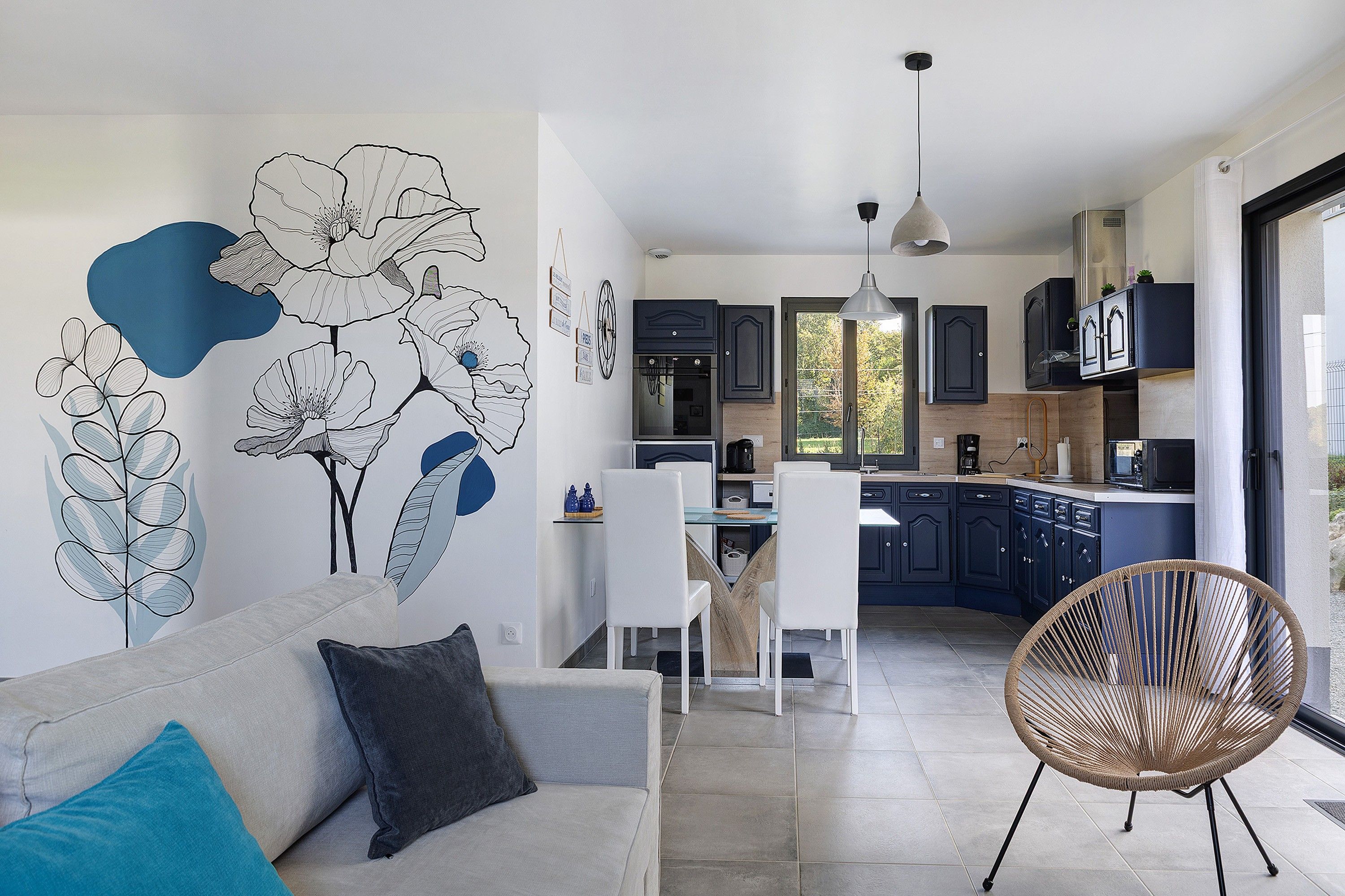 Une cuisine moderne avec des meubles de couleur sombre, un coin repas avec des chaises blanches et une grande fenêtre.