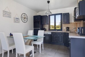 Een moderne keuken met donkere kastjes, een glazen eettafel, witte stoelen en een hanglamp.