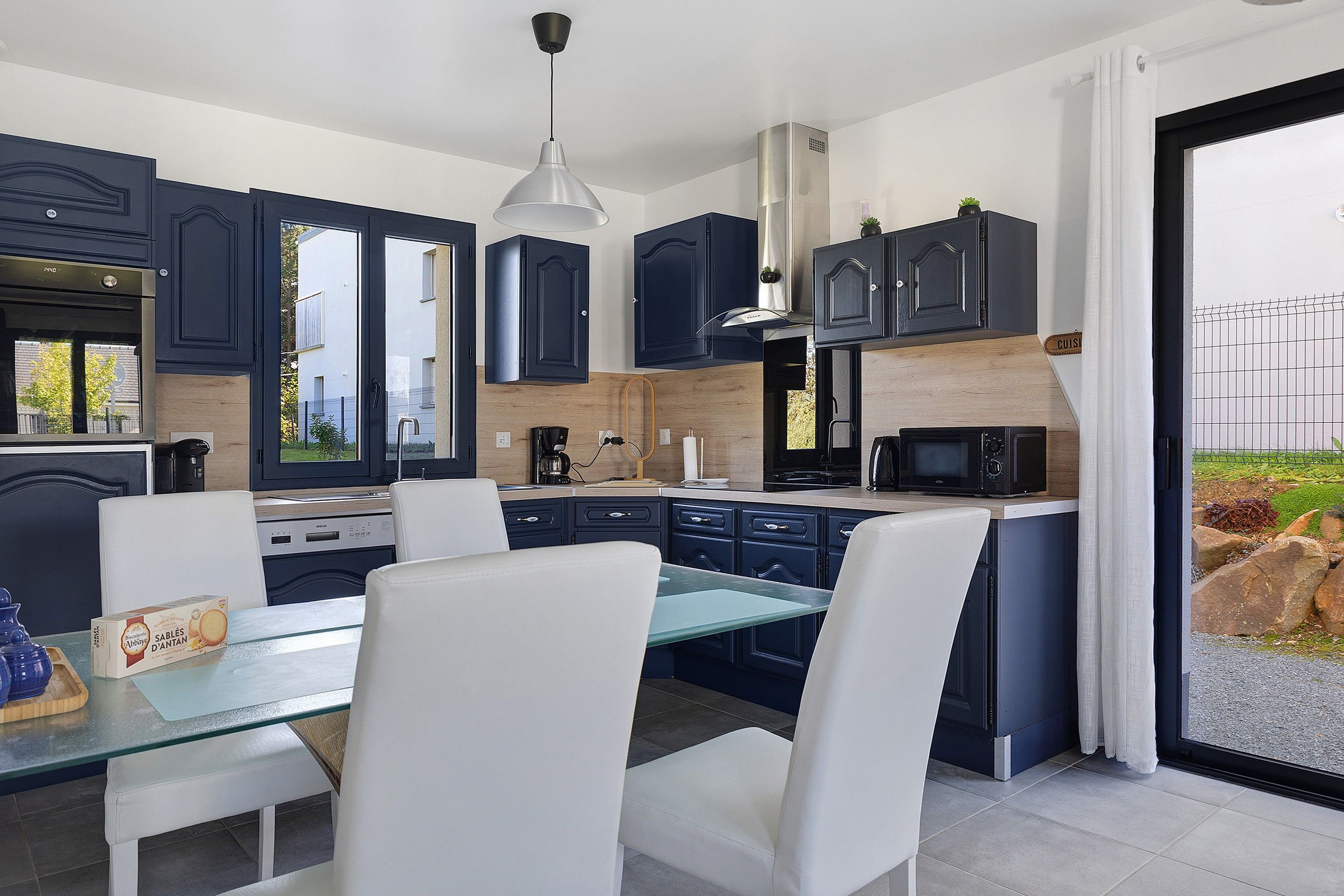 A modern kitchen with dark cabinetry, a dining table with white chairs, and a view of the outdoors through a glass door.