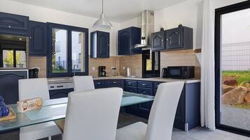 A modern kitchen with dark cabinetry, a dining table with white chairs, and a view of the outdoors through a glass door.