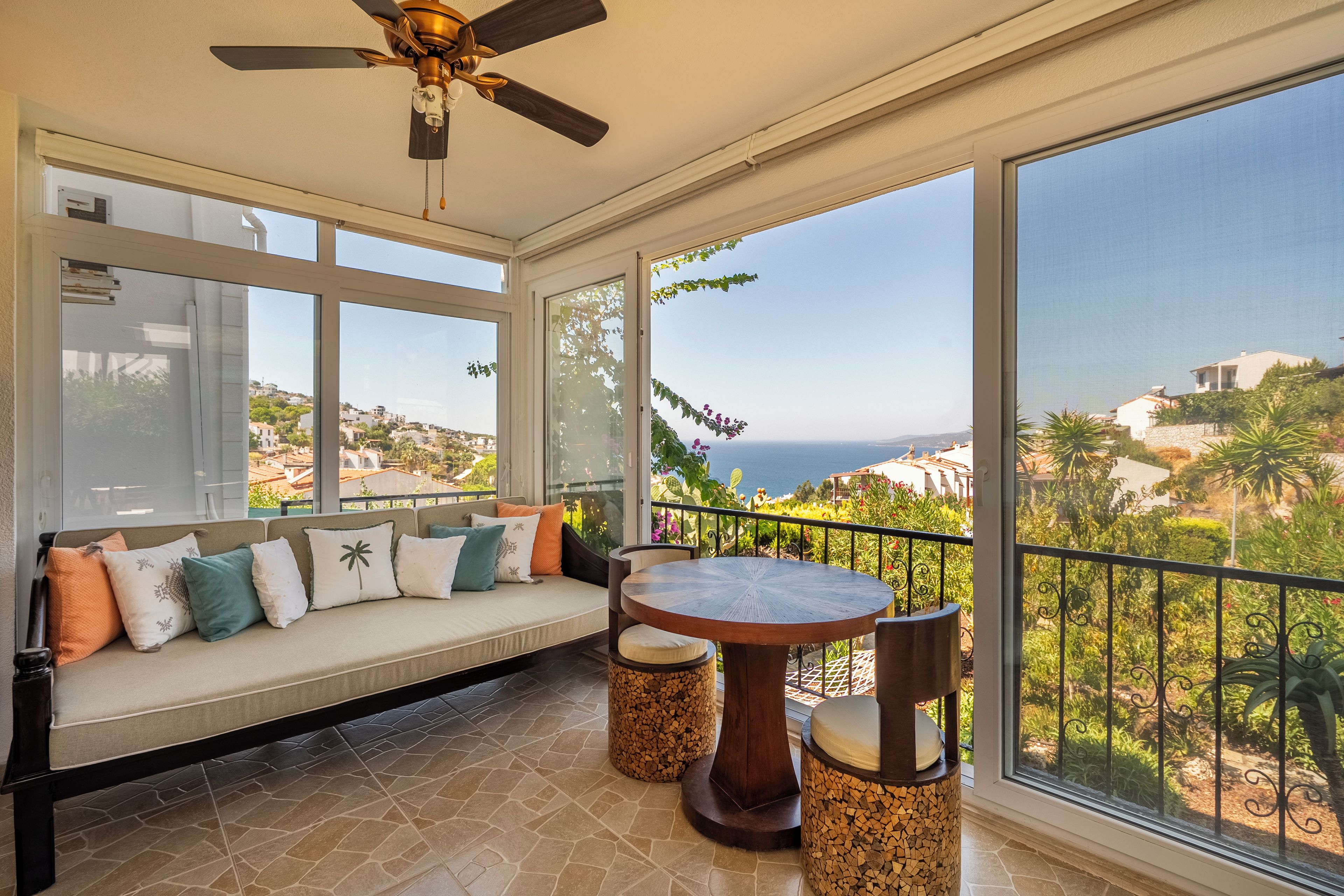 A sunlit balcony with a seating area, a table, and a view of the sea.