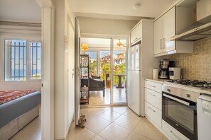 A modern kitchen with a built-in oven, a stove, and a refrigerator. There is a dining area with a table and chairs, and a view of the outside through a glass door. - Denize Sıfır Villa. Özel Plaj&Doğal Havuz.Bodrum (Milas)