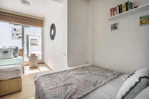 A bedroom with a bed, a bedside table, a dresser, a window with blinds, a wall-mounted light fixture, a door, and a shelf with books and a picture. - Denize Sıfır Villa. Özel Plaj&Doğal Havuz.Bodrum (Milas)