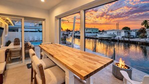 Un espace de restauration moderne avec une table et des chaises en bois, donnant sur une marina où sont amarrés des bateaux, et offrant une vue sur le coucher de soleil.
