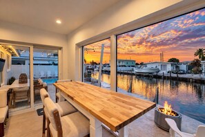 Uma sala de jantar moderna com mesa e cadeiras de madeira, com vista para uma marina com barcos e um pôr do sol.