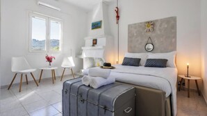 Habitació d'hotel amb un llit, una maleta, una taula petita amb flors, una cadira i una finestra amb vistes.