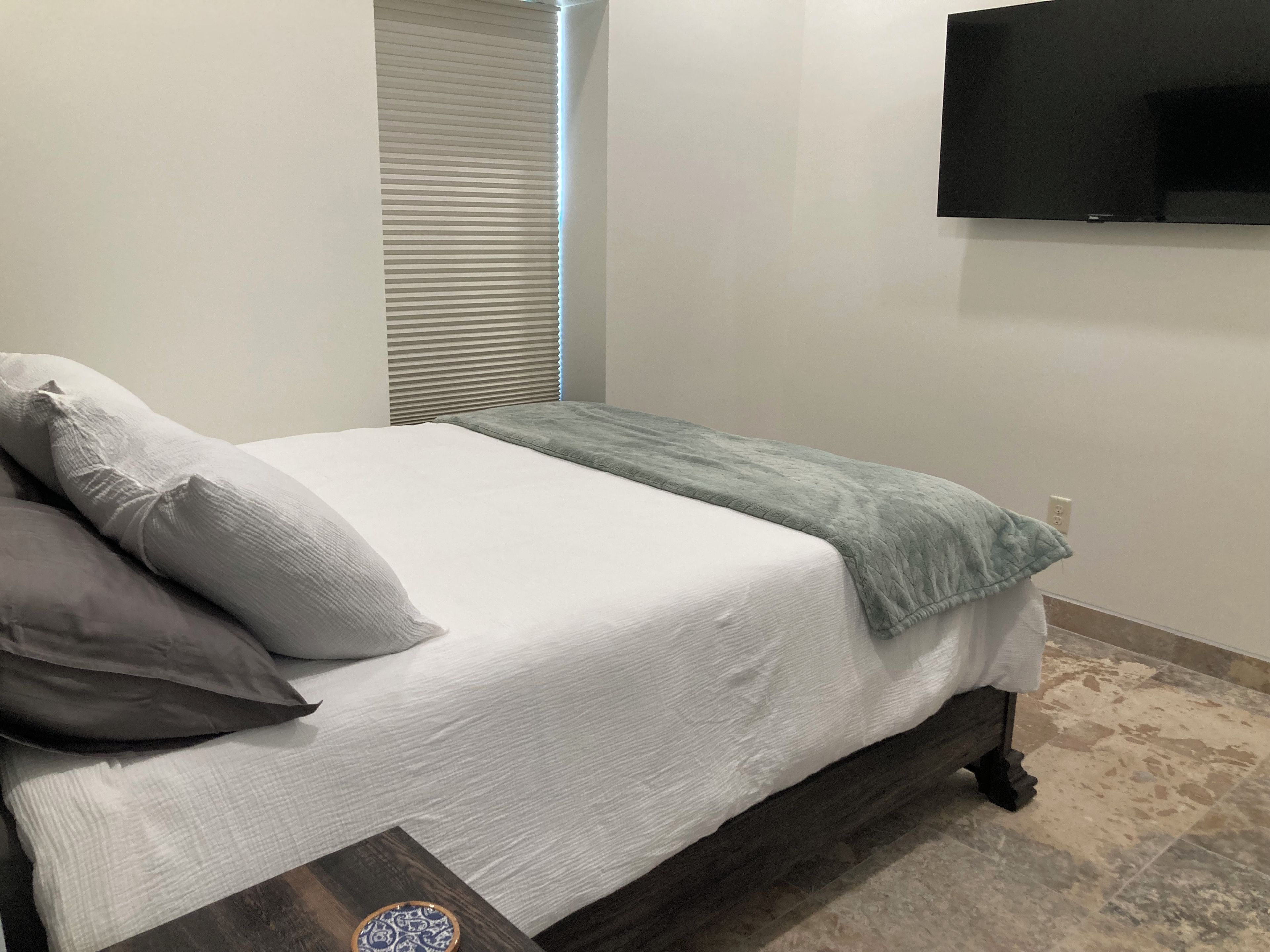 Ein ordentlich bezogenes Bett mit Kissen und Decke, ein Nachttisch mit einer Dekoration und ein an der Wand befestigtes Fernsehgerät.
