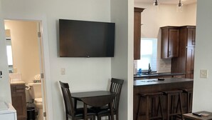 Un soggiorno moderno con un divano, un tavolo da pranzo con sedie, un angolo cottura con mobili in legno e un televisore a schermo piatto montato a parete.