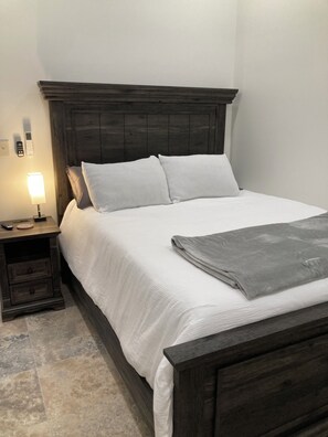 Una cama bien hecha con cabecera oscura, una lámpara de noche y una mesita de noche con cajones.