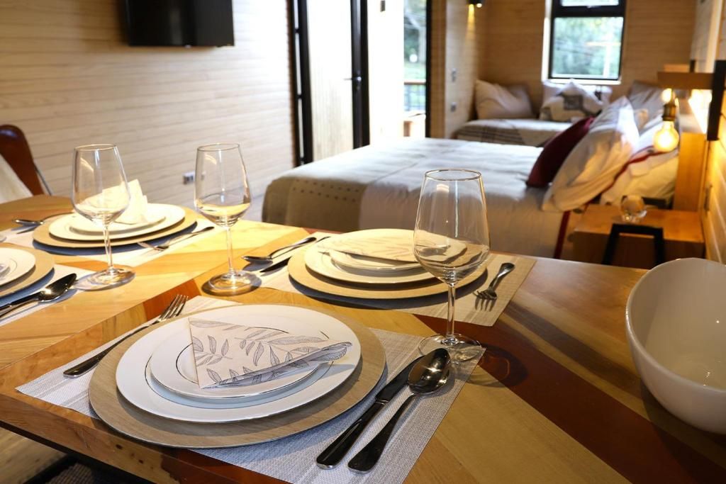 Una mesa de comedor bien puesta con platos, cubiertos y vasos, una cama al fondo y una mesita de noche con lámpara.