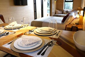 A neatly set dining table with plates, cutlery, and glasses, a bed in the background, and a bedside table with a lamp.