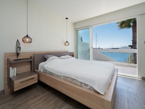 A modern bedroom with a large bed, wooden headboard, bedside tables, and a view of the sea through a sliding glass door.