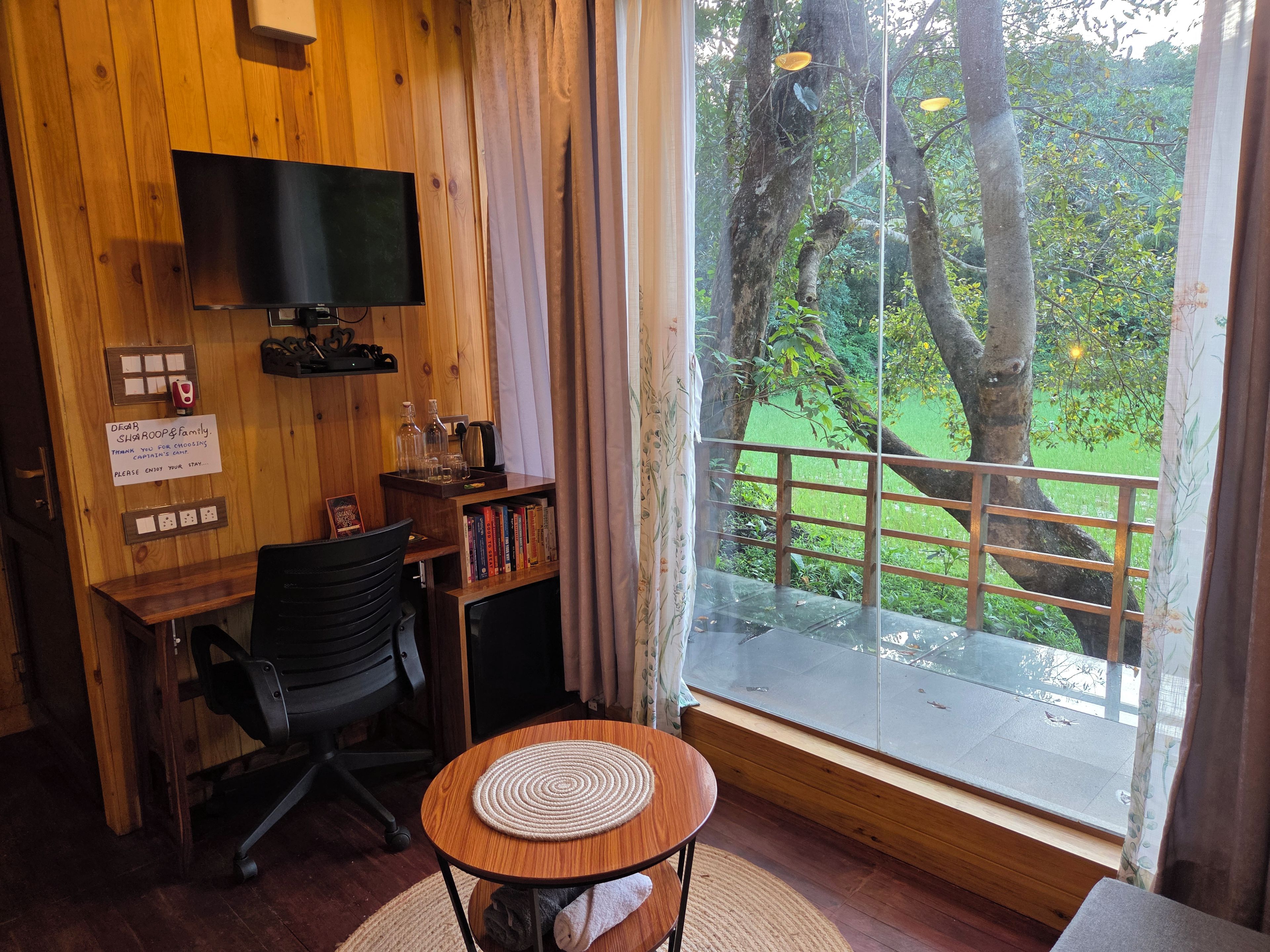 A room with a wooden desk, a chair, a television mounted on the wall, a bookshelf, and a window with a view of trees and a green field.
