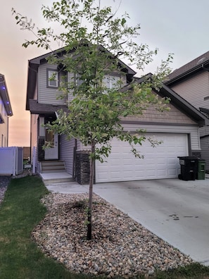 A residential house with a garage, a driveway, and a small tree in the front yard. - Room-charming and Cozy Home (Edmonton)