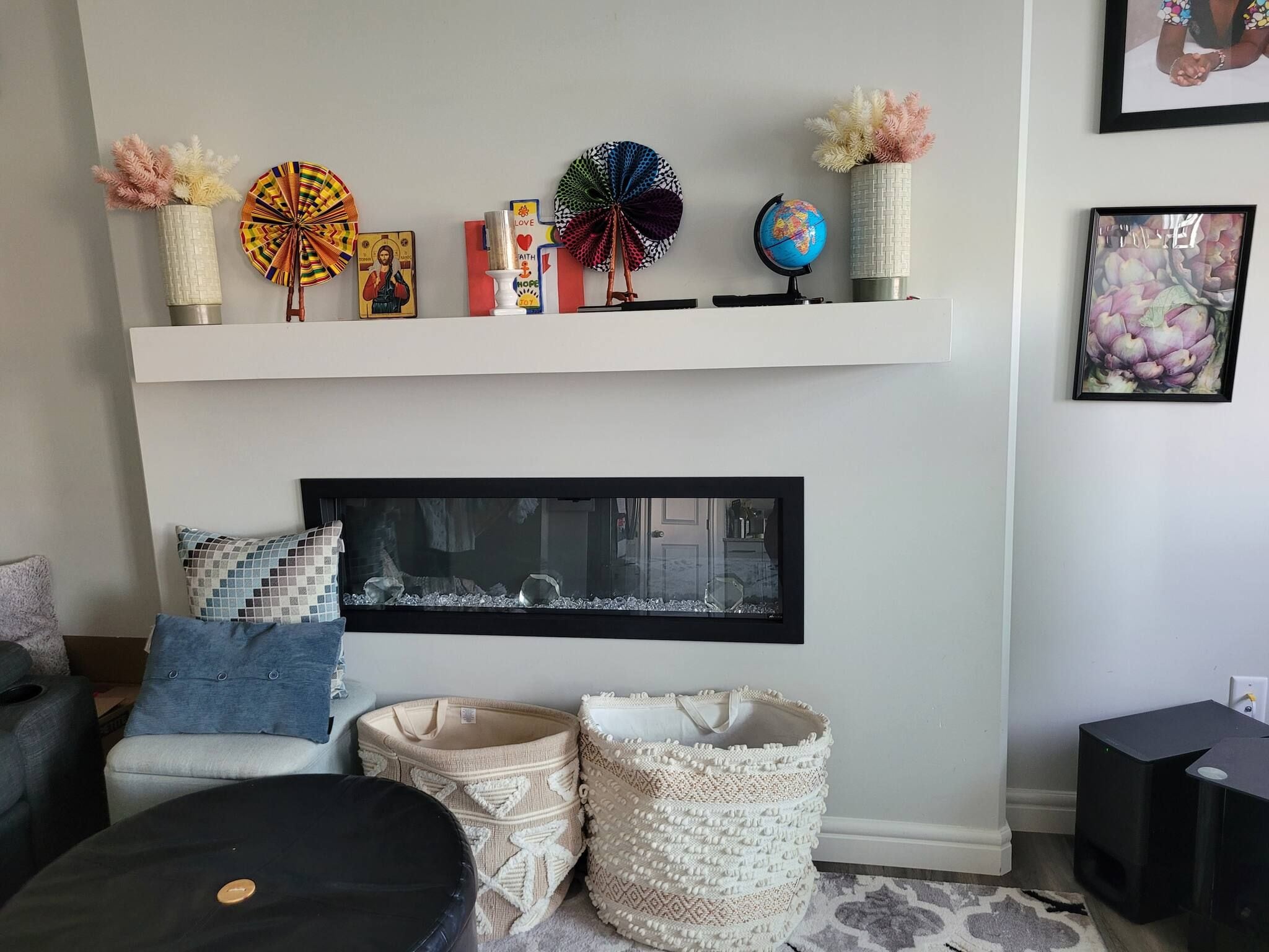 A living room with a fireplace, a shelf with decorative items, a framed picture, and a black ottoman.