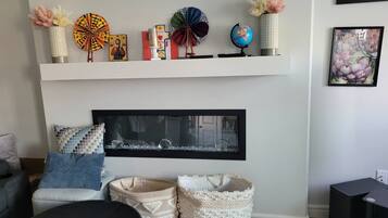 A living room with a fireplace, a shelf with decorative items, a framed picture, and a black ottoman.