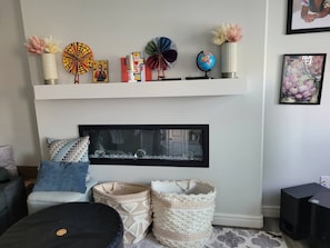 Een woonkamer met een open haard, een plank met decoratieve objecten, een ingelijste foto en een zwart voetenbankje.