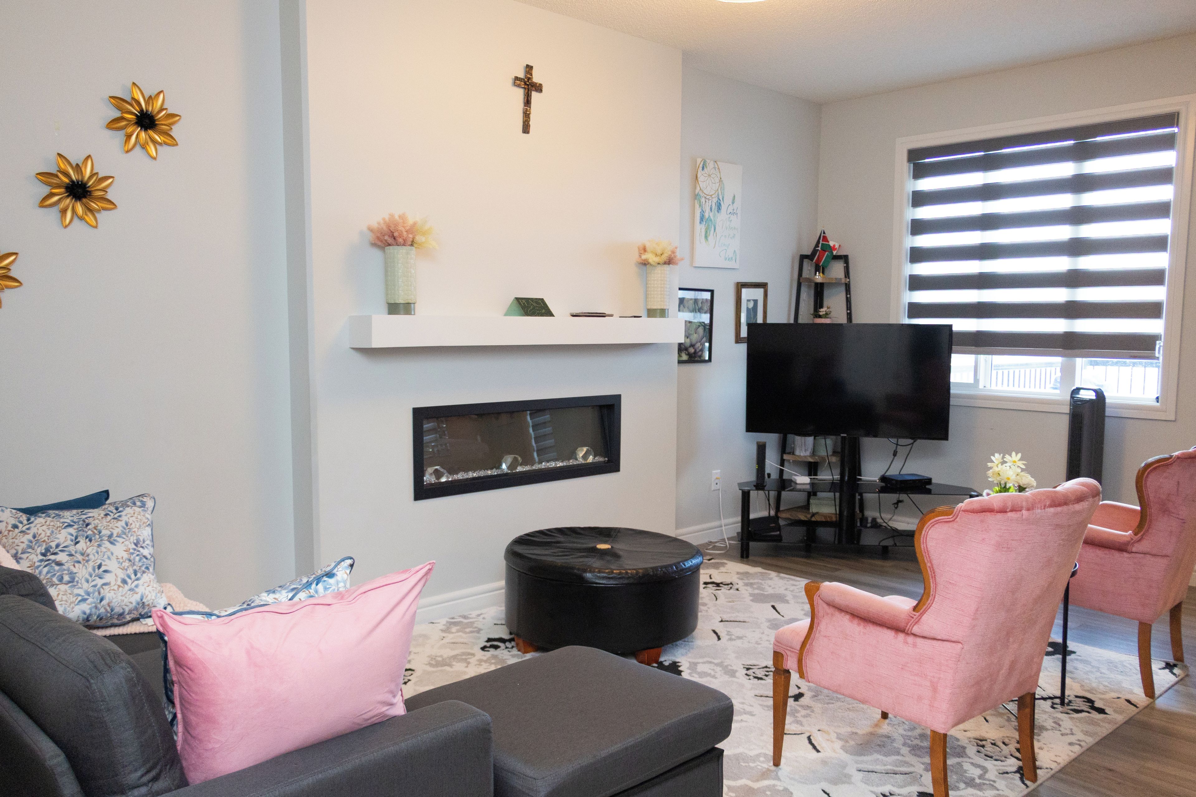 A living room with a sofa, ottoman, pink armchairs, a flat-screen TV, a fireplace, and decorative items on the wall.