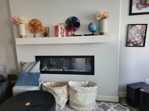 A living room with a fireplace, a shelf with decorative items, a framed picture, and a black ottoman. - Rapperswill Serenity (Edmonton)