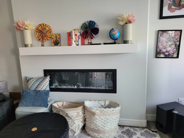 A living room with a fireplace, a shelf with decorative items, a framed picture, and a black ottoman. - Rapperswill Serenity (Edmonton)