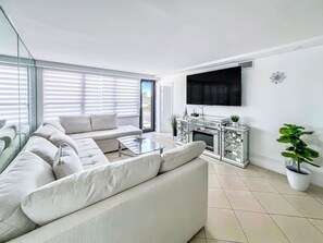 A modern living room with a sectional sofa, a glass coffee table, a fireplace with a decorative mantel, and a large window with blinds. - Charming 2BR/2BA Oceanfront Gem, Miami Beach (Miami Beach)