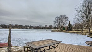 Una terraza de madera con una mesa y sillas de metal, con vistas a una masa de agua congelada y árboles y casas al fondo.