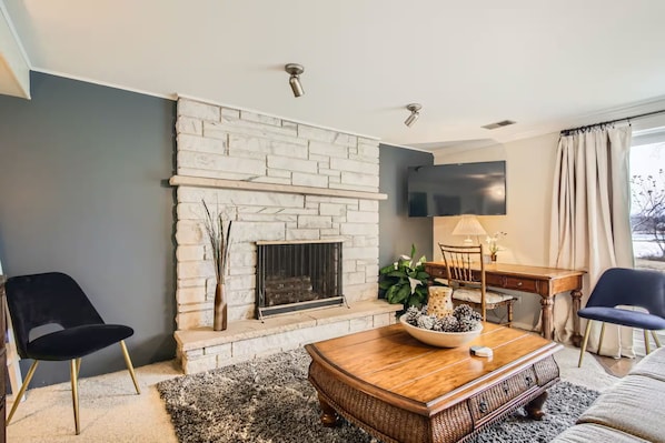 Una sala de estar con chimenea de piedra, un televisor de pantalla plana montado en la pared y un comedor con mesa y sillas de madera.