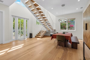 Ein modernes Interieur mit einer Holztreppe, einem Essbereich mit Tisch und Stühlen sowie einem großen Fenster mit Blick auf Grünflächen.