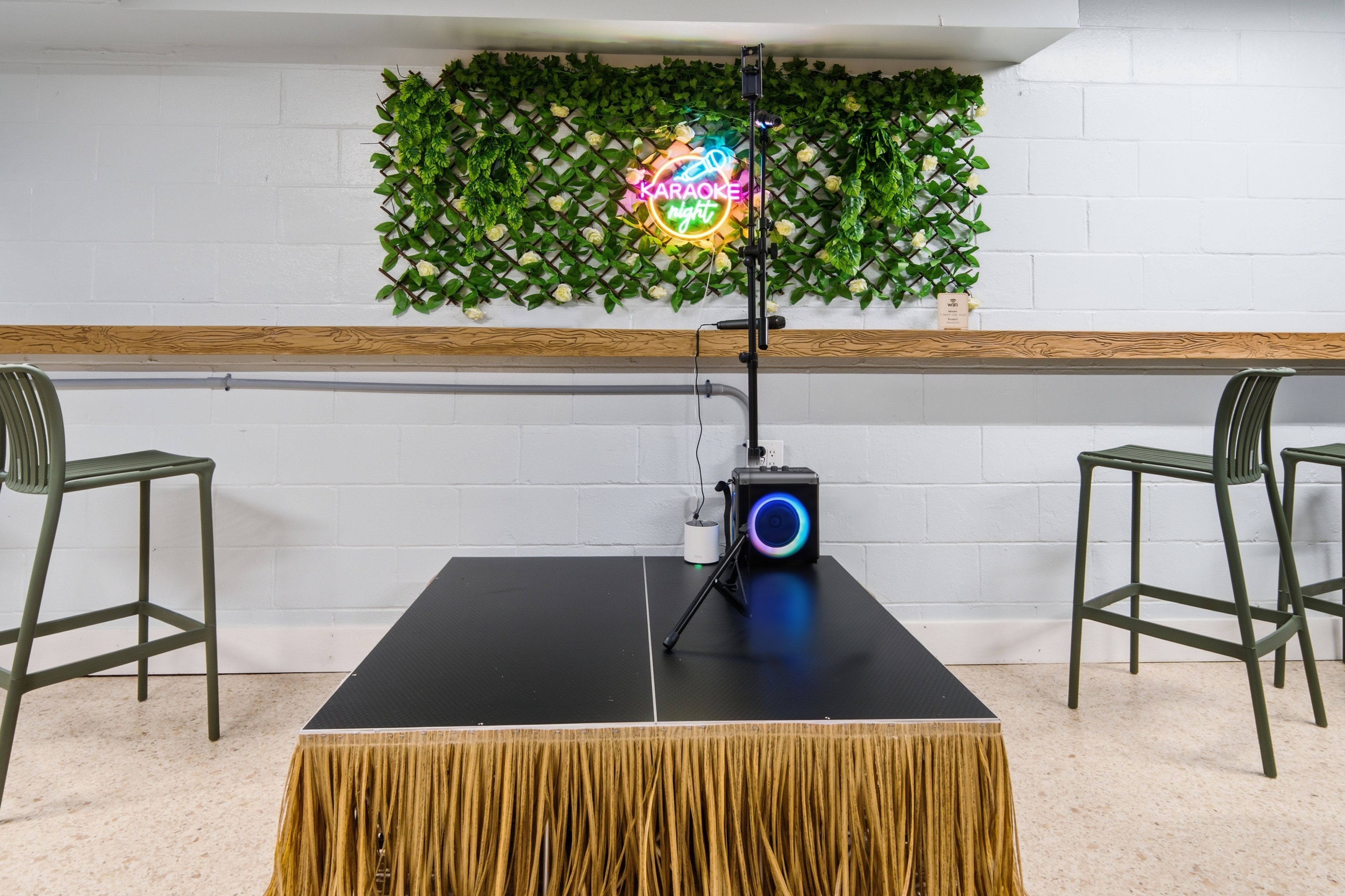 A room with a black table, two chairs, a neon sign, and a wall decorated with green foliage.