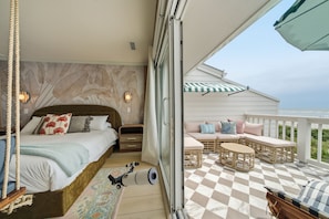 Un dormitorio con cama, mesitas de noche y un pequeño mueble auxiliar. Hay un balcón con mobiliario exterior y piso a cuadros.