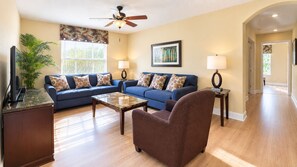 A living room with a blue sofa set, a coffee table, a brown armchair, a ceiling fan, and a television on a stand. - 6BR Family Vacation Home Paradise Palms Private Pool Near Disney (Kissimmee)