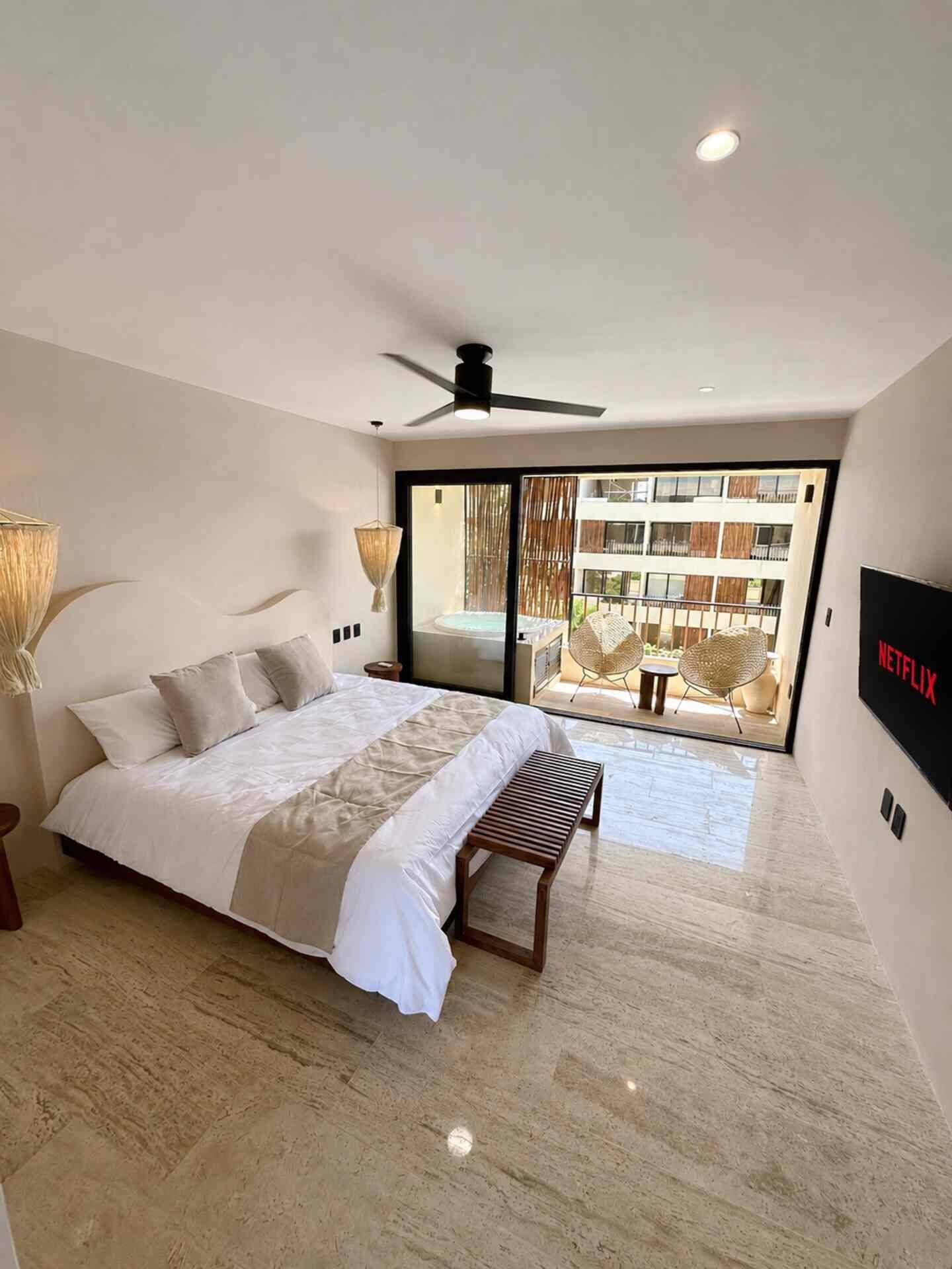 Une chambre moderne avec un grand lit, un ventilateur de plafond, une télévision et une vue sur un balcon aménagé avec du mobilier d’extérieur.