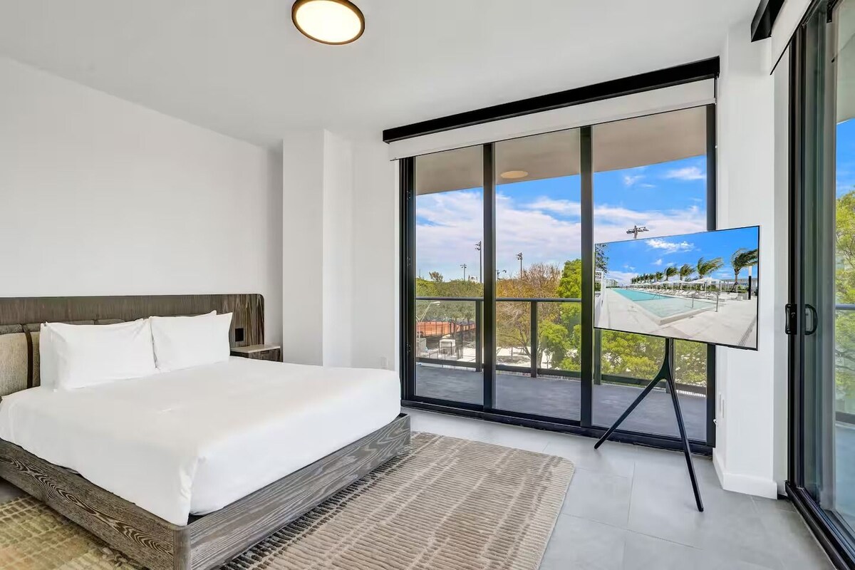 A modern bedroom with a large bed, a TV on a stand, and a balcony with a pool view.