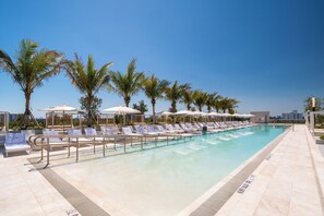 Área de piscina com espreguiçadeiras e guarda-sóis, palmeiras e céu claro.