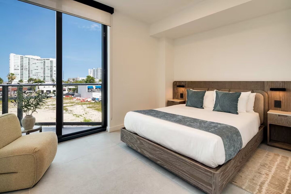 A modern hotel room with a large bed, a sofa, a view of the city, and a balcony.