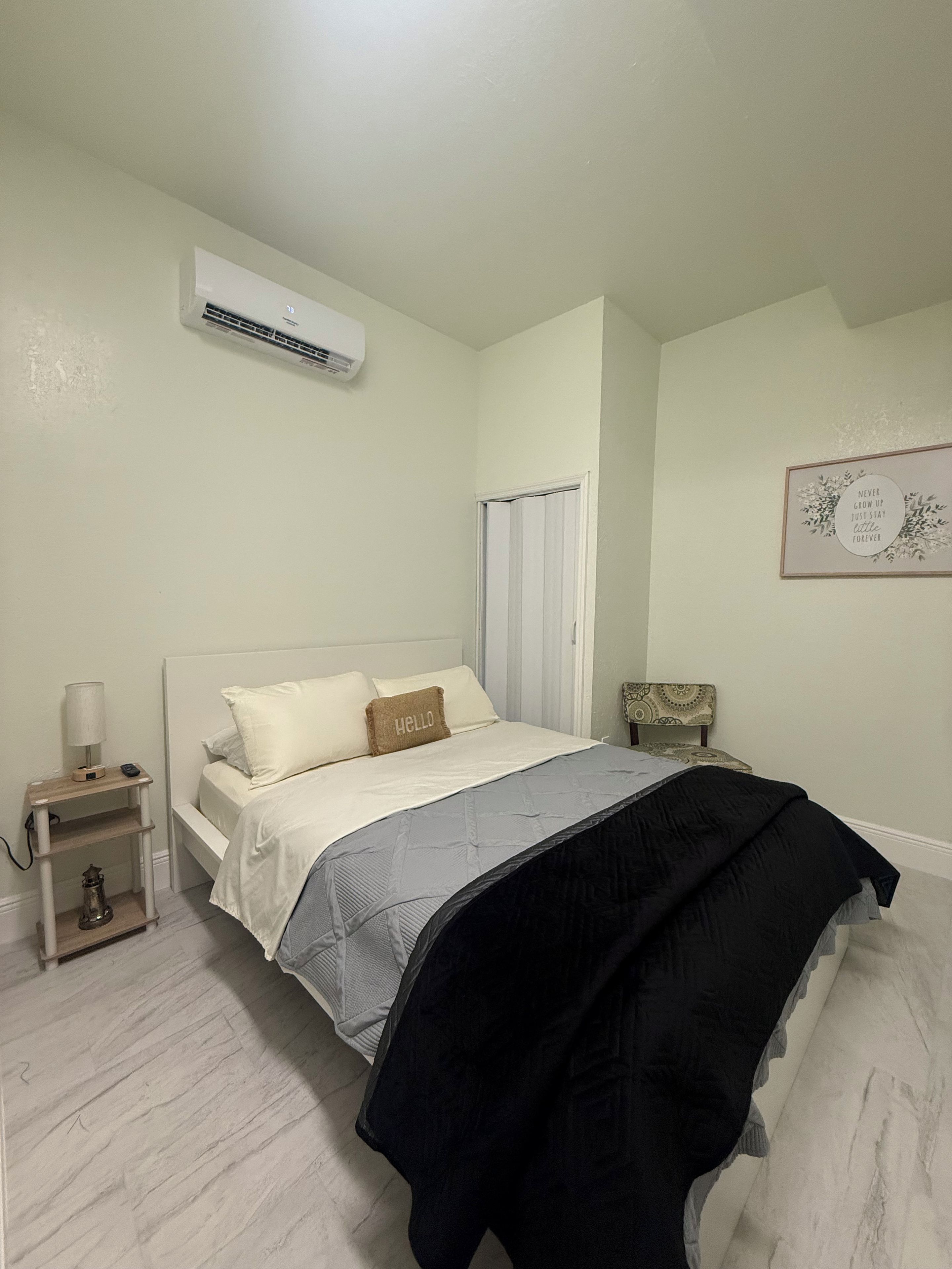 A bedroom with a bed, bedside table, lamp, and a wall-mounted air conditioner.