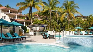 Piscina con sillas de descanso y sombrillas, rodeada de palmeras y edificios de hotel.