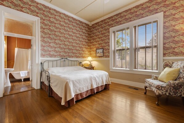 A bedroom with a bed, a chair, a window with blinds, and a bathroom with a shower curtain.